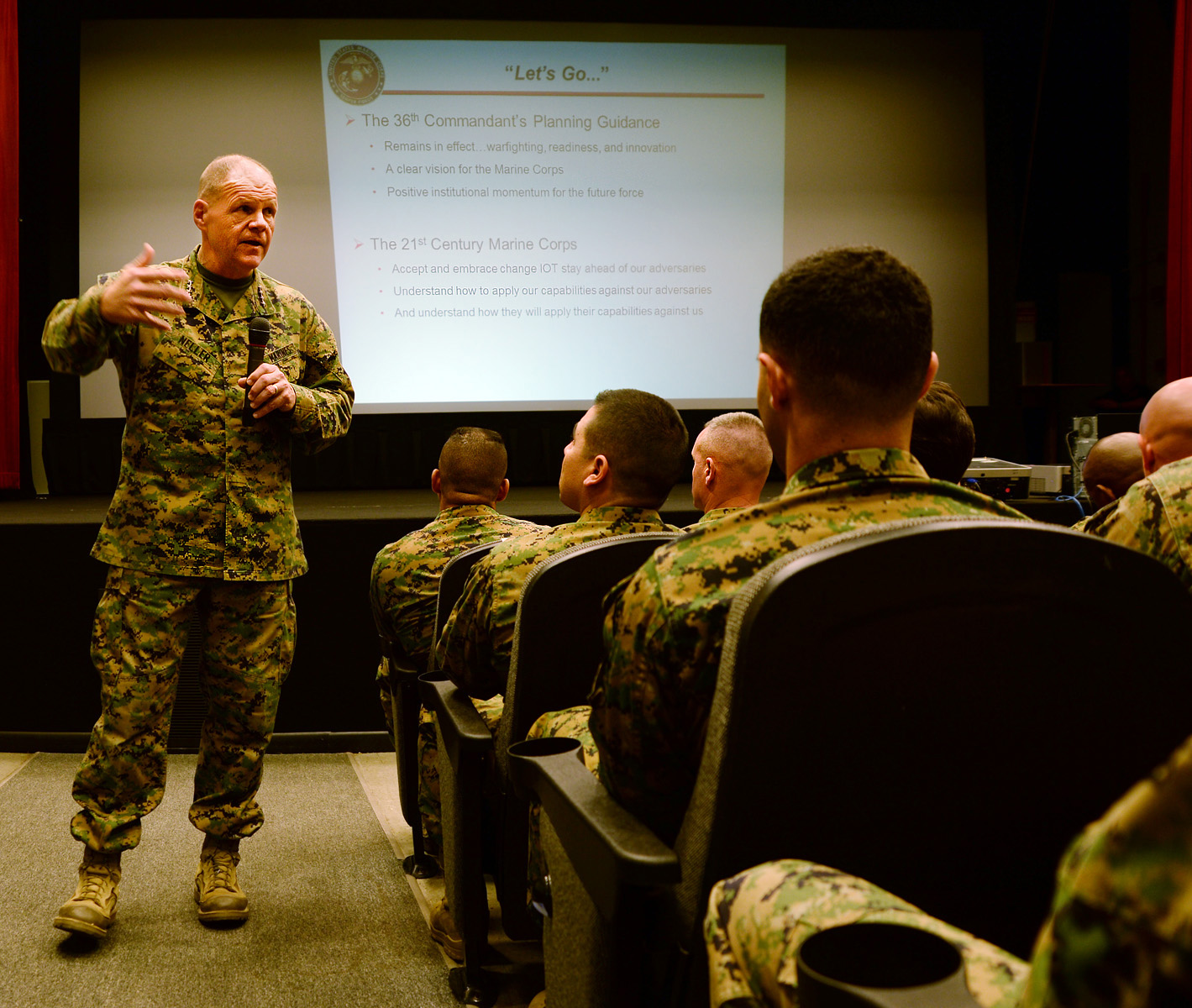 A Marine Colonel in digital camouflage uniform briefs junior officers in a command briefing room, with a presentation screen behind showing strategic planning guidance and institutional momentum.