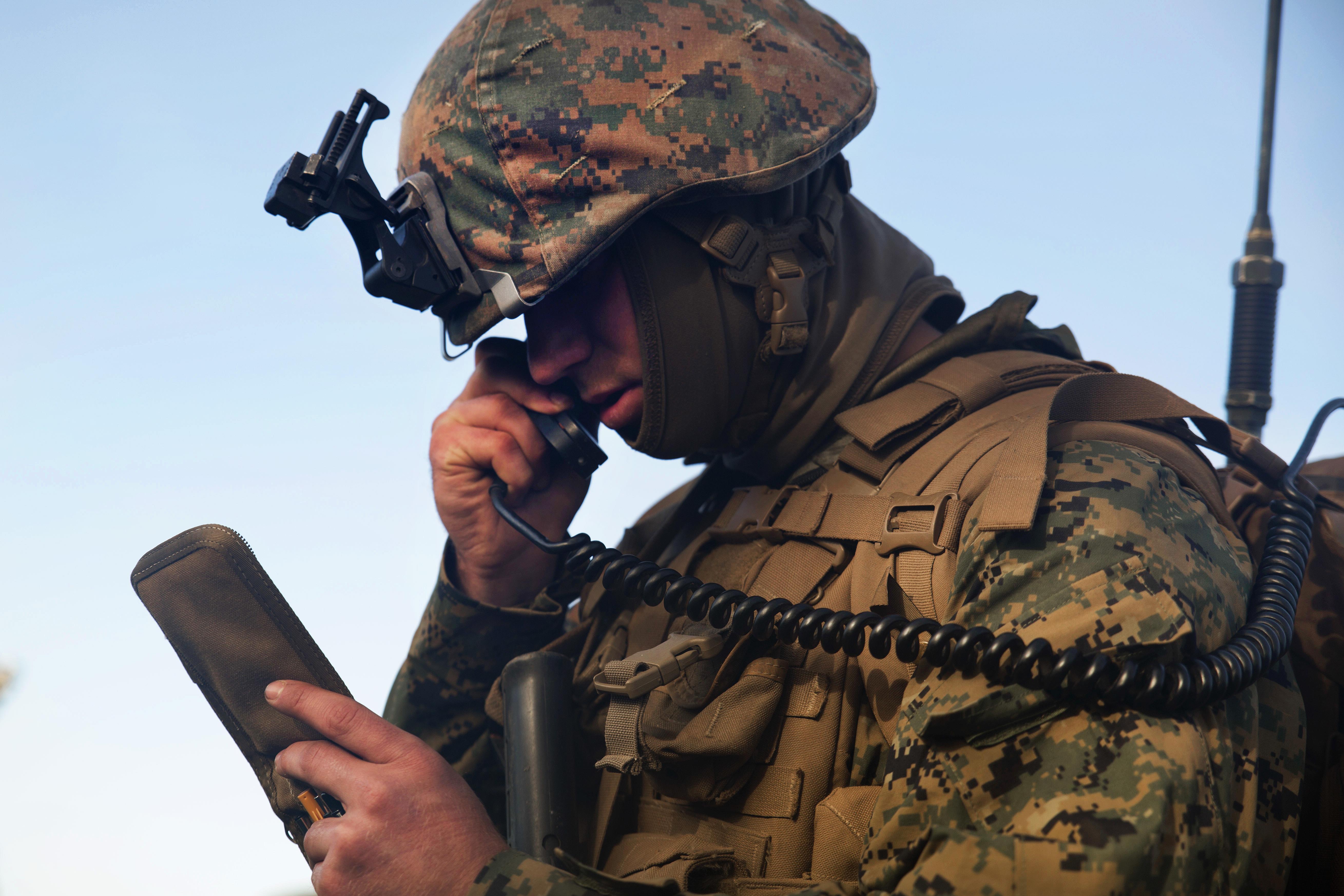 A Marine squad leader in digital camouflage uniform and helmet communicates on a tactical radio handset, checking coordinates on a printed map, exemplifying real-time command and unit direction.